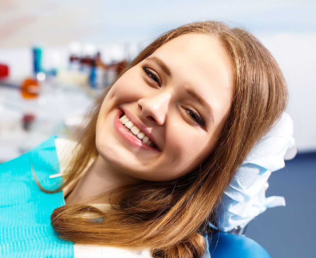 A young woman with long, light brown hair smiles brightly while reclining in a dental chair, wearing a blue dental bib. The background shows blurred dental equipment.