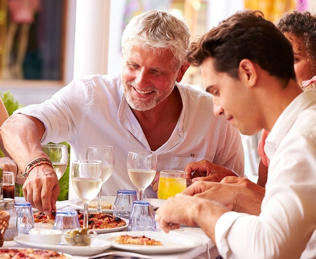 Two men sit outdoors at a table, smiling and enjoying a meal with pizza and drinks. There are several glasses and plates on the table, and other people are partially visible in the background.