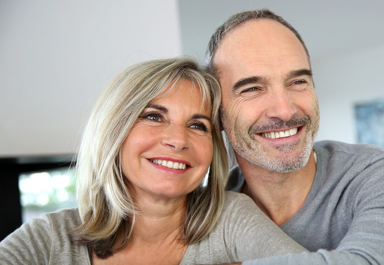 A smiling middle-aged couple with gray hair sits close together, looking content and happy in a bright indoor setting.