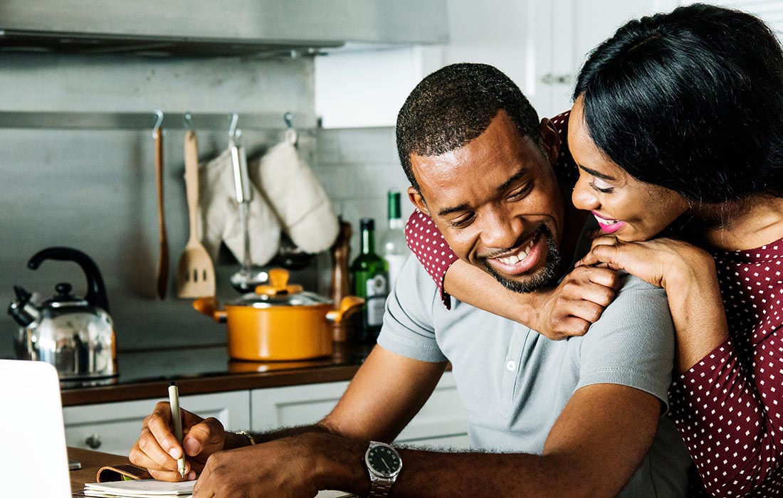 A smiling woman hugs a man from behind as he writes on paper at a kitchen table. Both are happy, surrounded by kitchenware and a laptop, in a cozy, modern kitchen.
