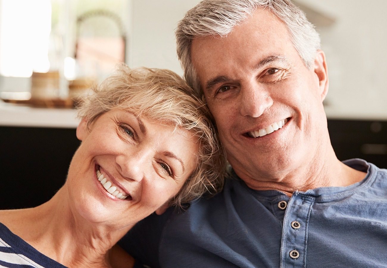 Smiling older couple sitting close together indoors, both looking at the camera. The woman has short blonde hair, and the man has short gray hair. They appear happy and relaxed.