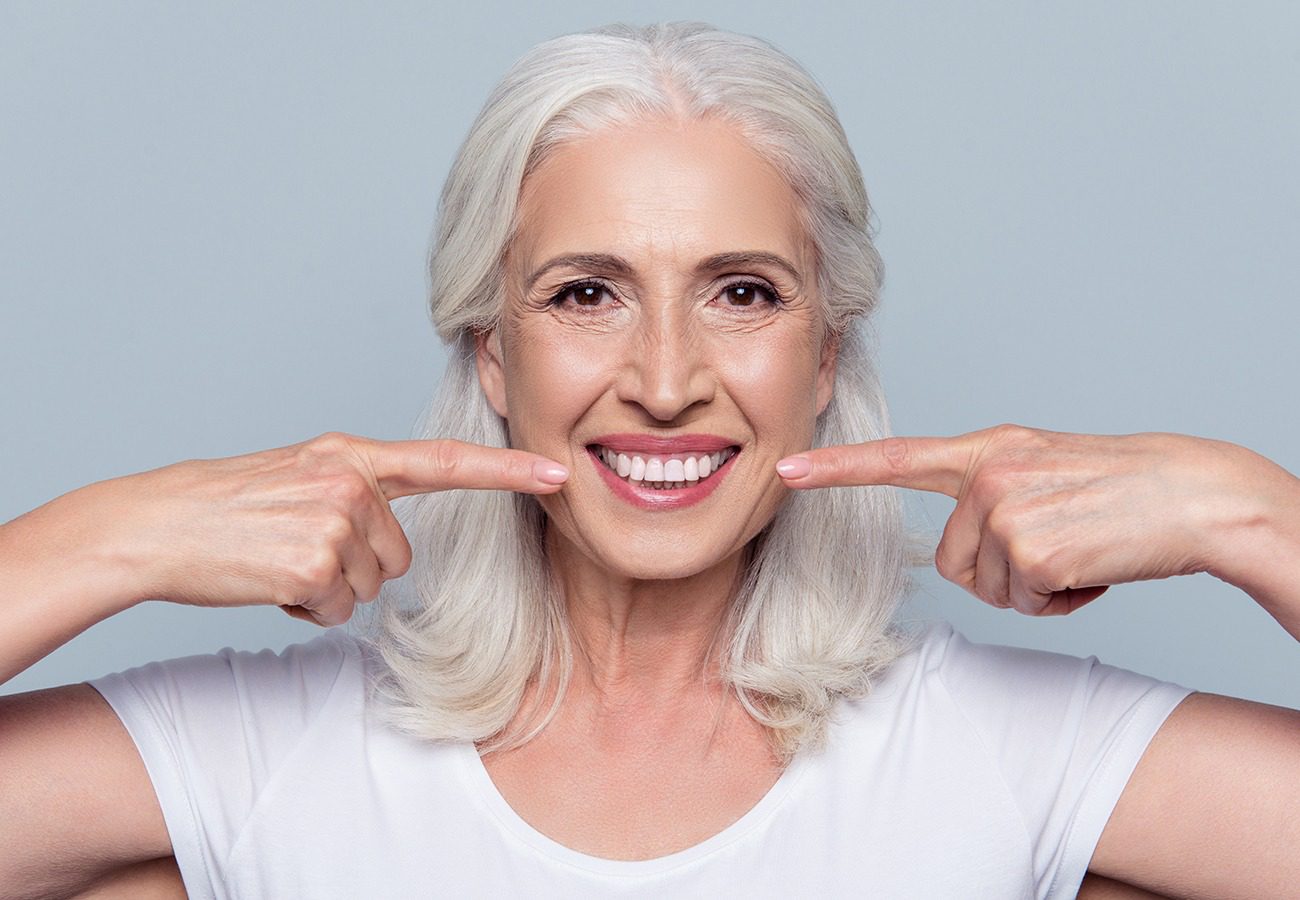 Smiling older woman with long gray hair wearing a white shirt, pointing to her teeth with both index fingers against a plain light background.