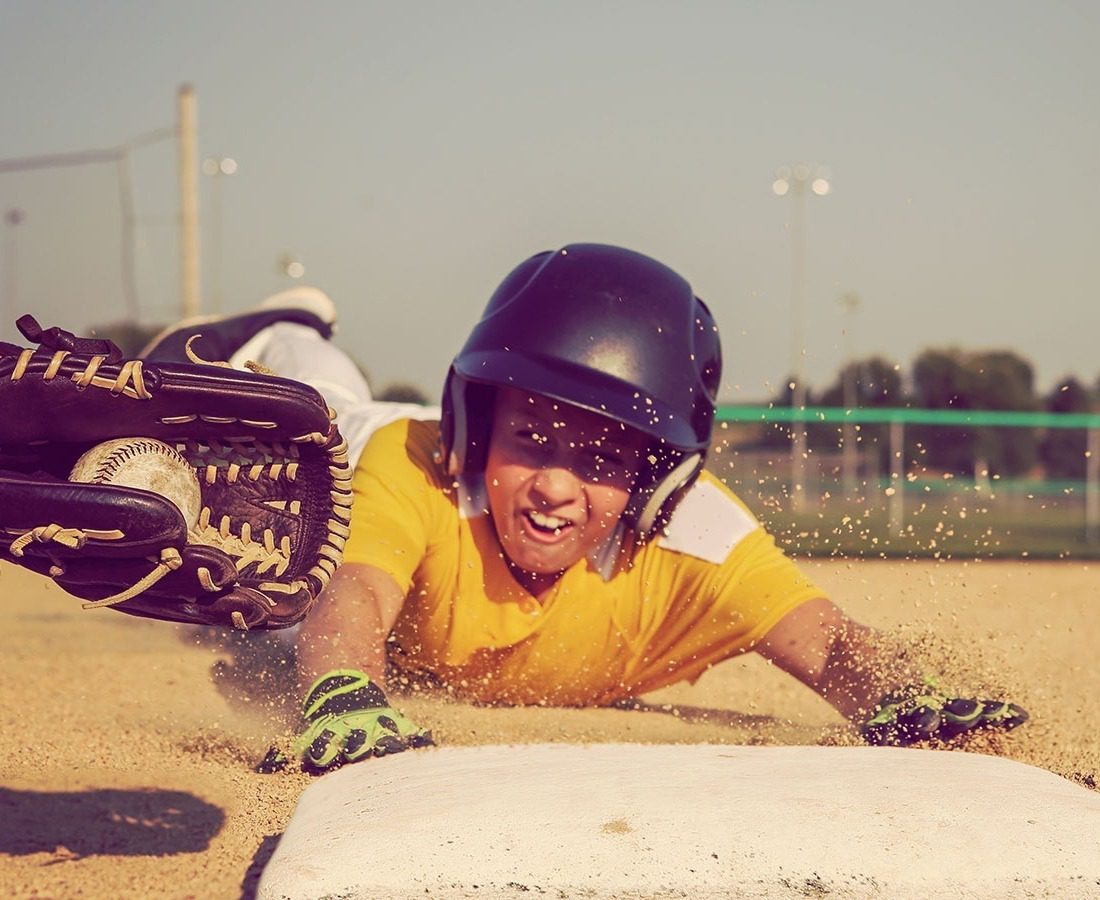 A young baseball player in a yellow jersey and helmet slides headfirst into a base as a gloved opponent attempts to tag him, kicking up sand on a sunny day at an outdoor field.