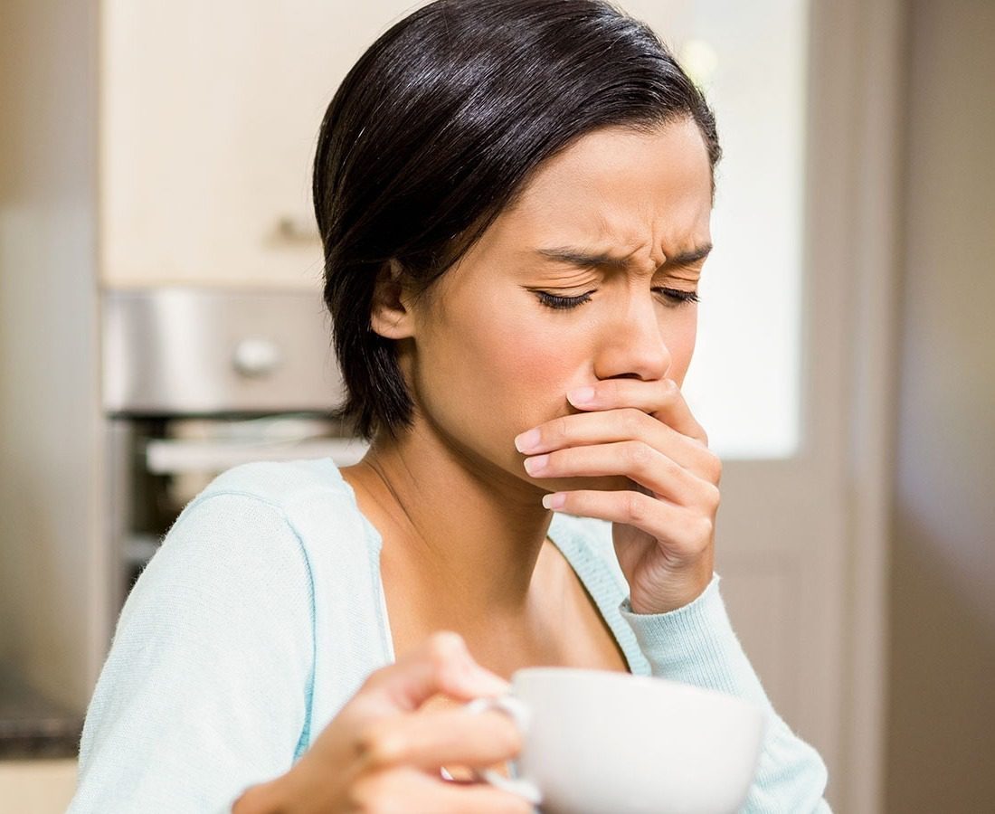 A woman with short dark hair holds a cup in one hand and covers her mouth with the other, appearing to feel nauseous or unwell, while sitting indoors in a kitchen.