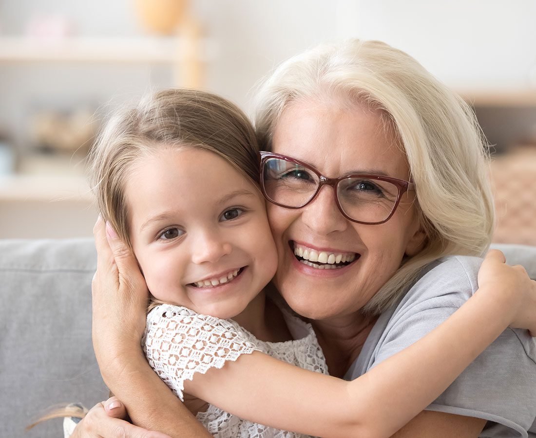 An older woman with glasses and gray hair hugs a young girl with long brown hair. Both are smiling and looking at the camera, sitting close together on a couch in a bright room.