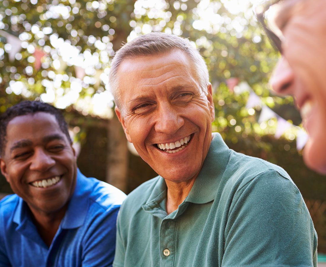 Three people sit outdoors, smiling and laughing together. The focus is on a middle-aged man in a green shirt, with two others—one in blue and one in glasses—enjoying the moment in a sunlit, leafy setting.