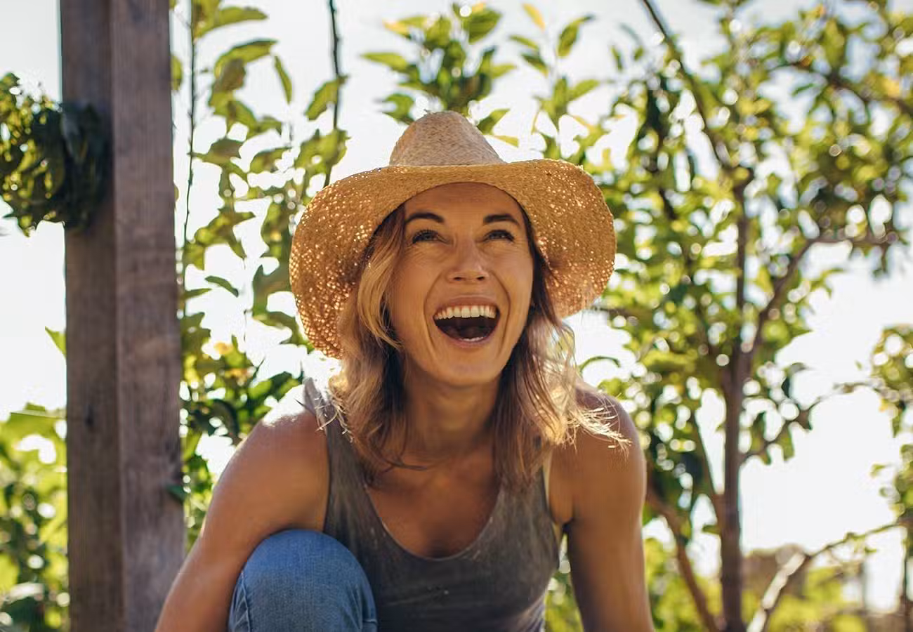 A woman wearing a straw hat and tank top smiles and laughs outdoors, surrounded by green leafy plants and sunshine.