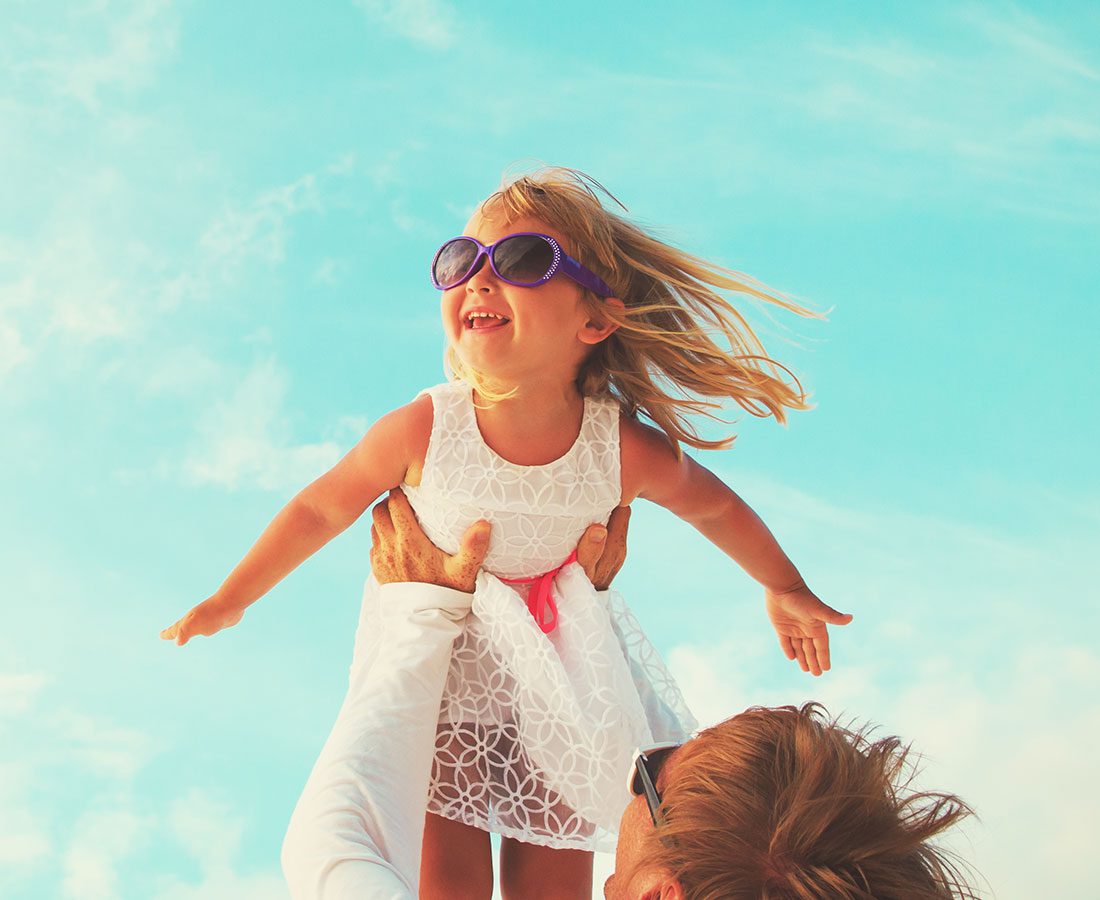 An adult lifts a smiling young girl in a white dress and sunglasses into the air against a bright blue sky. The girl’s arms are outstretched and her hair blows in the wind.