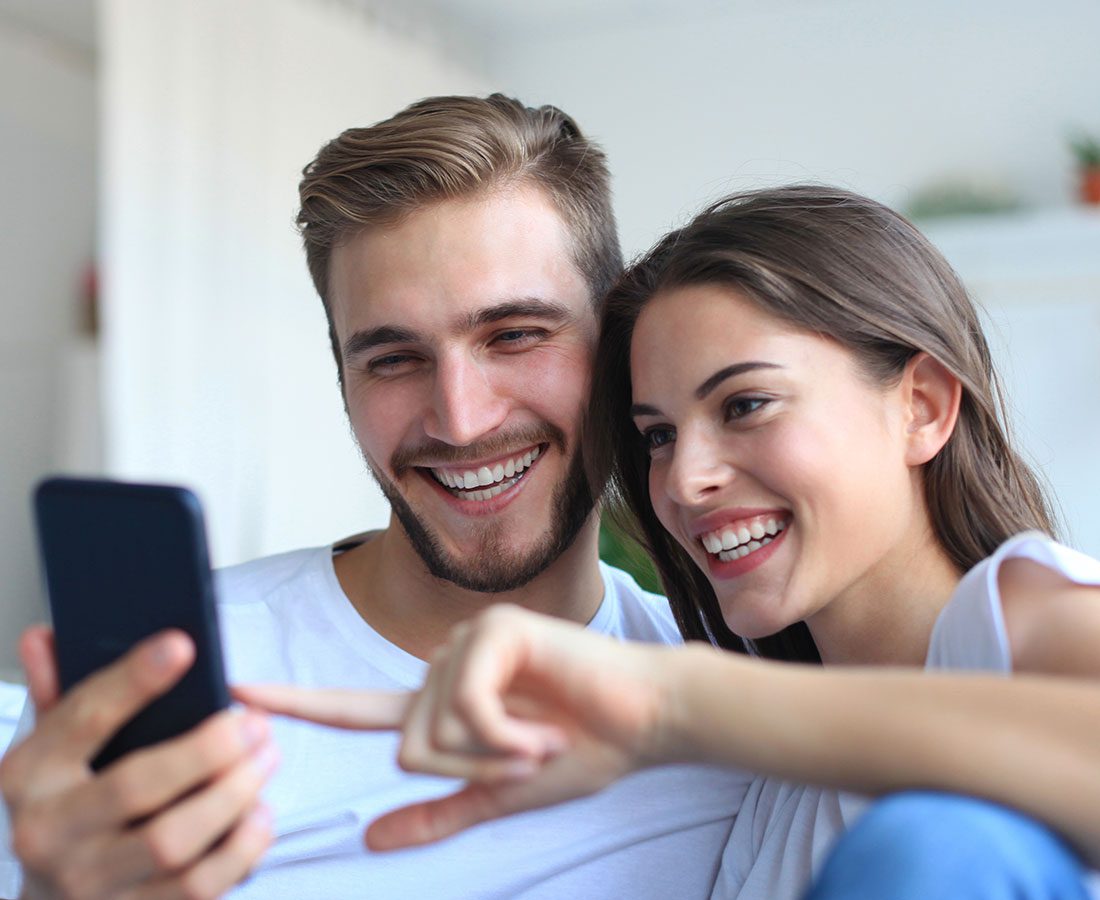 A smiling man and woman sit close together, looking at a smartphone. The woman points at the screen, and both appear happy and engaged. They are indoors, wearing casual white shirts.