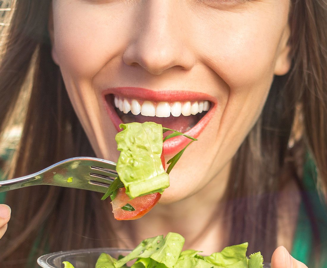 A smiling woman holds a fork with lettuce and tomato close to her mouth, about to eat a fresh salad. Her teeth are visible, and she appears happy and healthy.