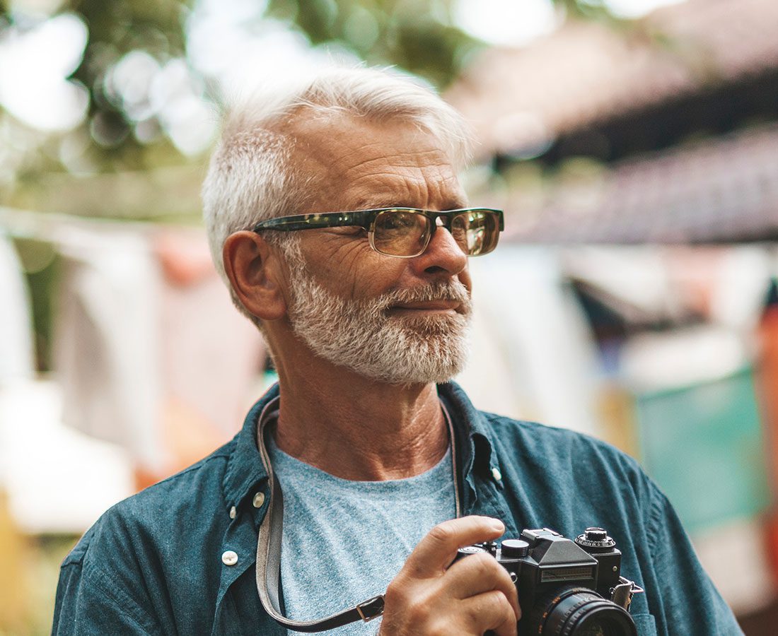 An older man with gray hair, a beard, and glasses holds a camera outdoors, wearing a blue shirt over a light t-shirt. The background is blurred with hints of trees and buildings.