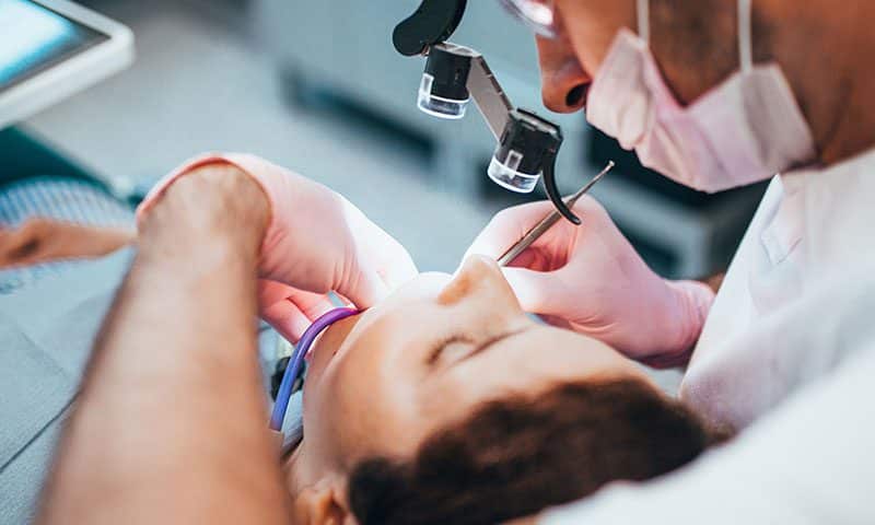A dentist wearing magnifying glasses and pink gloves examines a patients mouth with dental tools while the patient lies back in the dental chair.