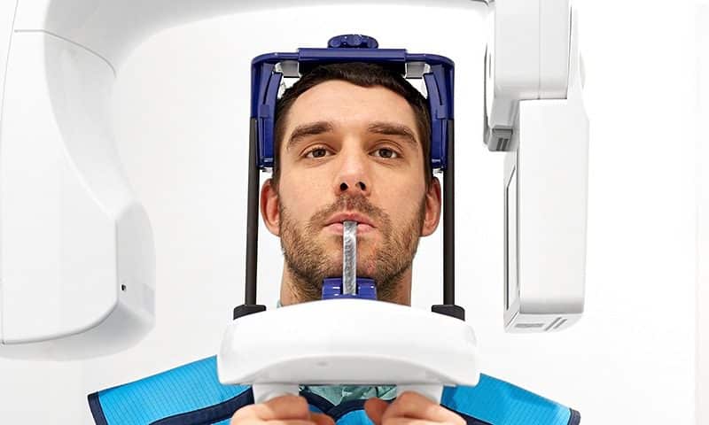 A man sits with his head positioned in a dental X-ray machine, biting a plastic piece and wearing a protective lead apron, preparing for a panoramic dental scan.