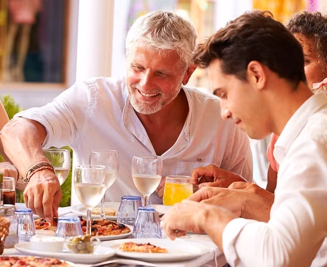 Two men are enjoying a meal together at an outdoor restaurant, smiling and sharing food. There are plates of pizza, glasses of white wine, and orange juice on the table. Other people are partially visible in the background.