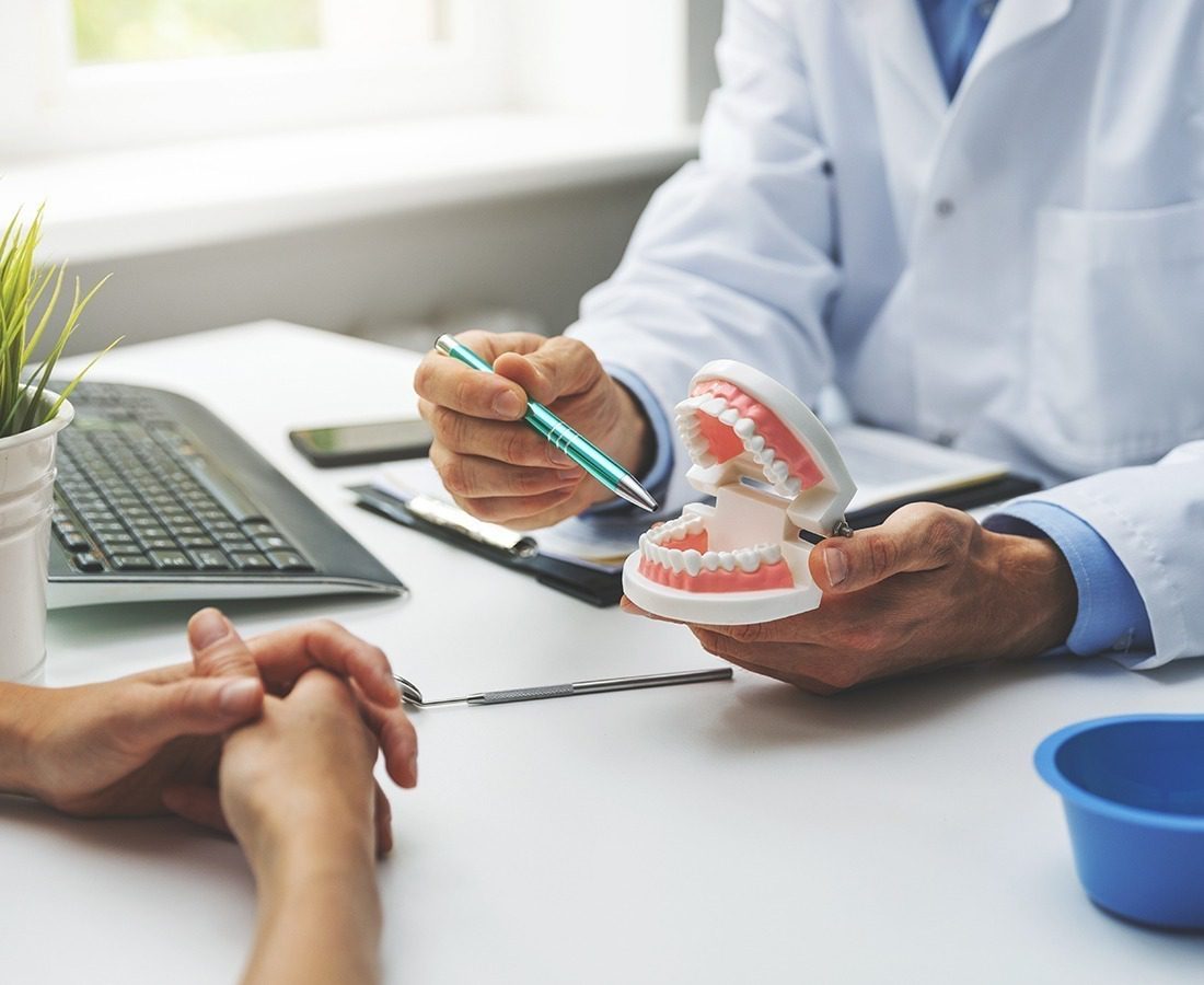 A dentist in a white coat points at a dental model with a pen, explaining something to a patient sitting across the desk. A keyboard, dental tools, and a small plant are also visible on the desk.