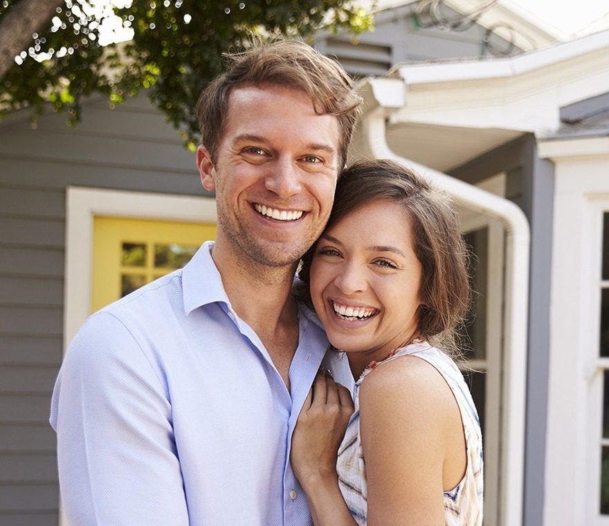 A smiling couple stands close together outside a house with gray siding and a yellow door, looking happy and relaxed in the daylight.