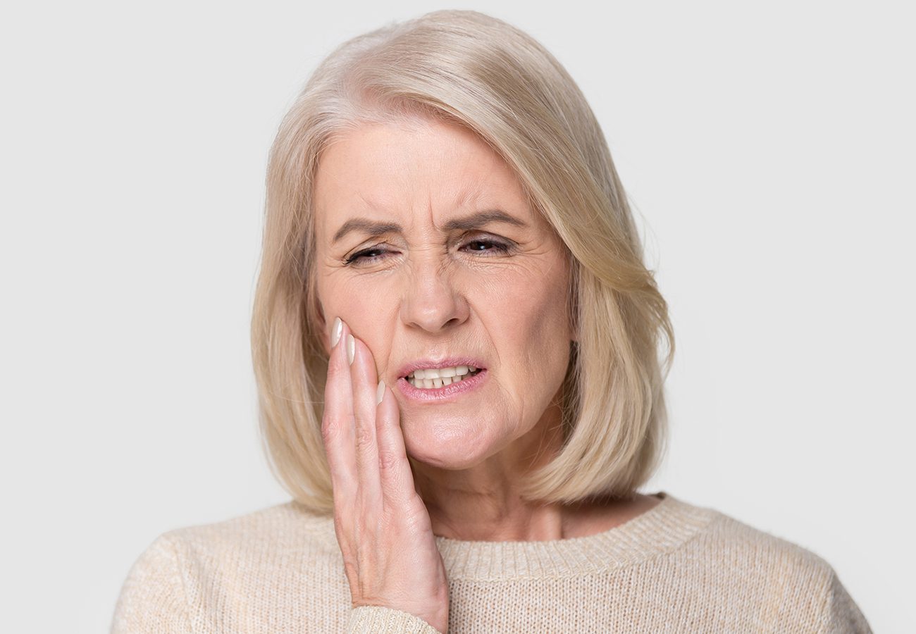 Older woman with short blonde hair touching her cheek and wincing in pain, possibly experiencing toothache or discomfort, against a plain light background.