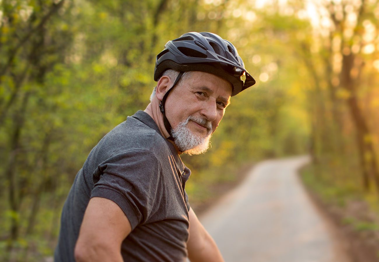 An older man with a gray beard, wearing a black bicycle helmet and dark shirt, smiles while sitting on a bike on a tree-lined path during a sunny day.