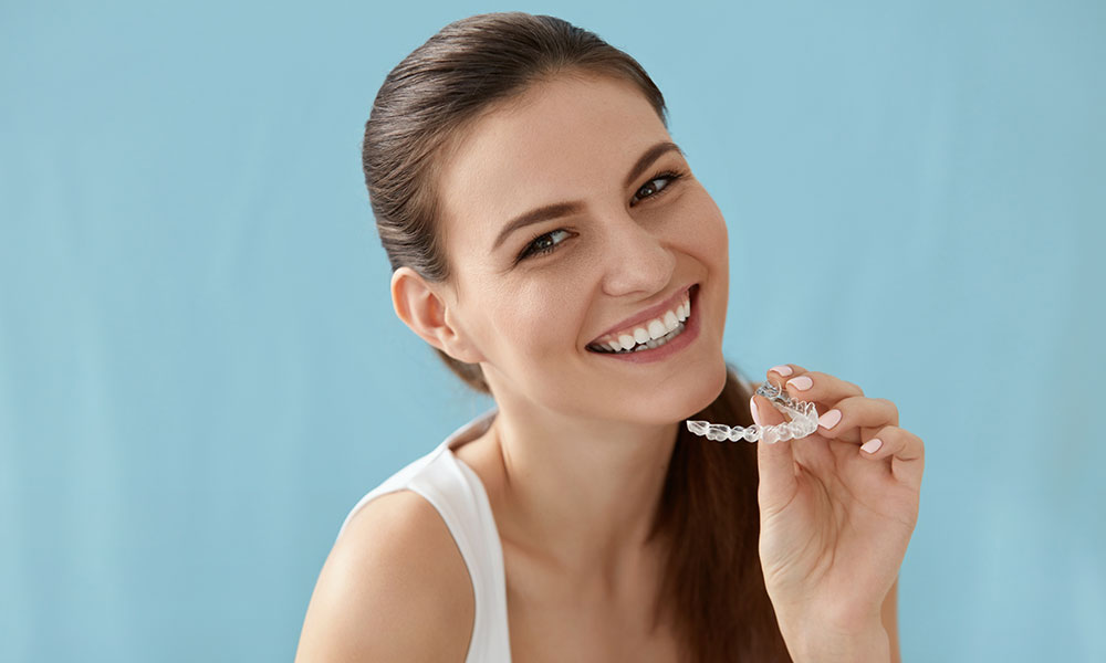 Smiling woman with brown hair holds a clear dental aligner in her hand against a light blue background.